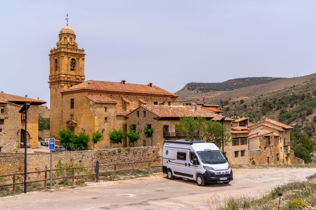 Descubre la libertad de viajar en camper. Imagen de una furgoneta camper aparcada junto a un pintoresco pueblo medieval rodeado de montañas.